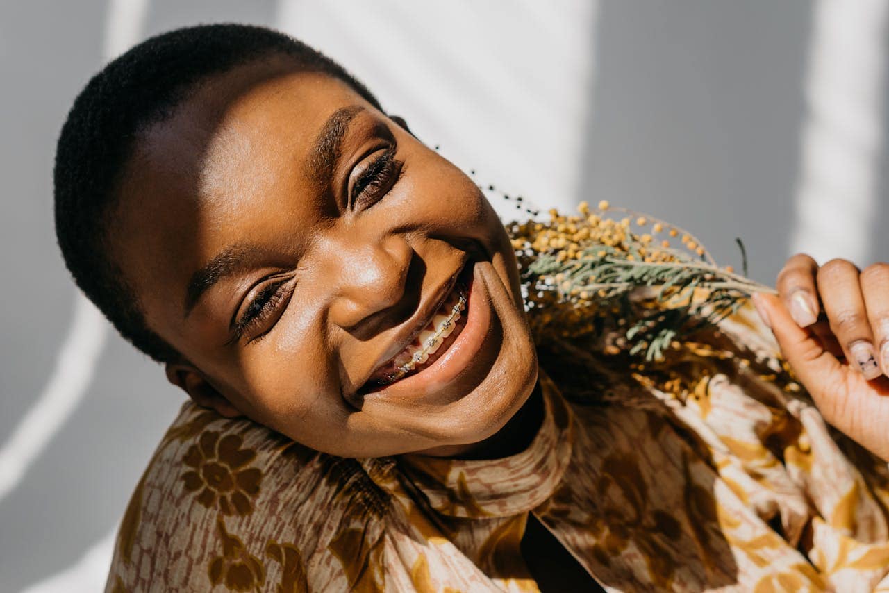 Smiling woman with braces tilting her head, holding a sprig of yellow mimosa flowers in sunlight